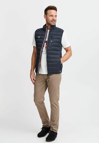 Navy puffer vest with vertical seams, high collar, and zip pockets, worn over a white graphic t-shirt, paired with beige pants and black shoes.