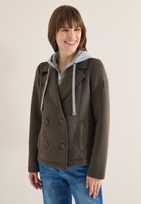Femme portant un manteau croisé vert olive foncé superposé à un sweat à capuche gris zippé avec des cordons, associé à un jean bleu, souriant légèrement vers la gauche.