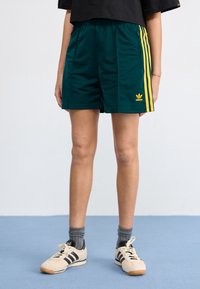 Gröna sportshorts med gula ränder, elastisk midja och en Adidas-logotyp. Kombineras med krämfärgade skor och grå strumpor.