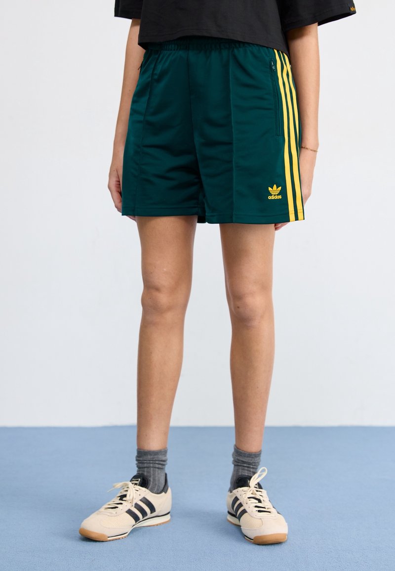 Gröna sportshorts med gula ränder, elastisk midja och en Adidas-logotyp. Kombineras med krämfärgade skor och grå strumpor.