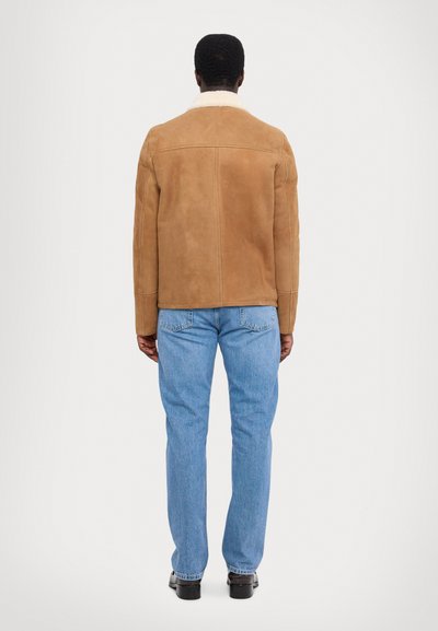 Tan ruskindsjakke med hvidt shearling-krave, tætsiddende design, lige snit og lange ærmer, iført lyseblå jeans og sorte sko.
