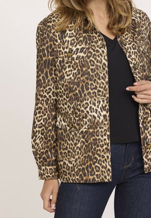 Frau trägt eine leo-bedruckte Knopfjacke über einem schwarzen Shirt und dunkelblauen Jeans, steht mit einer Hand nahe der Jacke.