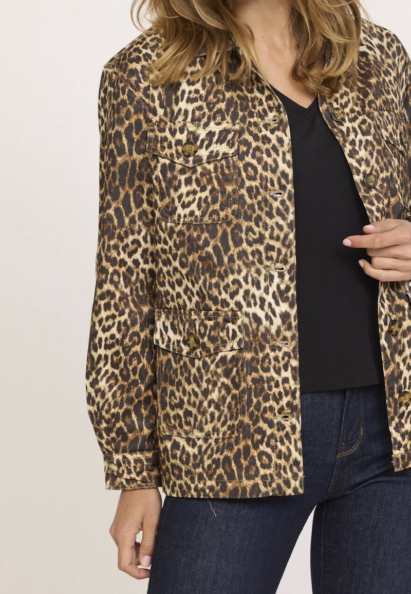 Femme portant une veste à boutons imprimé léopard par-dessus un chemisier noir et un jean bleu foncé, debout avec une main près de la veste.