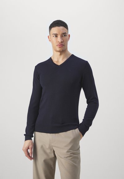Pull en maille bleu marine avec un décolleté en V, manches longues, poignets et ourlet côtelés, associé à un pantalon couleur crème. Texture douce, design ajusté.