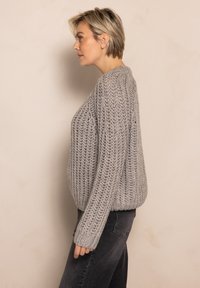 Knitted gray sweater with a loose fit, textured pattern, and wide sleeves, paired with dark jeans. Side view against a neutral background.