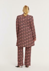 Patroonpak in zwart, roze en rood met ovale motieven; lange jas en wijde broek; gladde stof; zichtbare stilettohakken.