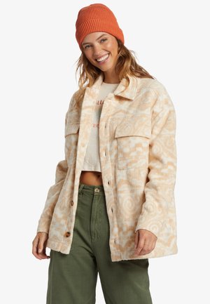Fleece shirt jacket in light beige with abstract swirl pattern, featuring two large front pockets and button closure. Paired with olive green pants.