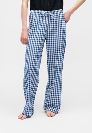 Personne portant un pantalon de pyjama à cordon bleu et blanc à carreaux amples, debout pieds nus sur un fond uni.