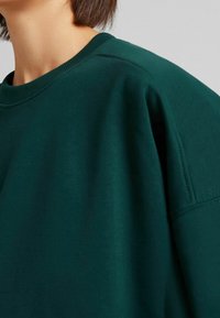 Sudadera verde oscura con cuello redondo, que presenta una textura suave y costuras de hombro caídas. La costura es visible a lo largo de los bordes.