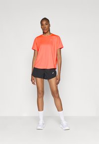 Γυναίκα που στέκεται με φωτεινό πορτοκαλί κοντομάνικο πουκάμισο, μαύρο αθλητικό σορτς, λευκές κάλτσες και λευκά αθλητικά παπούτσια σε απλό φόντο.