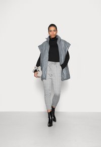 Grijze puffer vest met hoge kraag, gedragen over een zwarte coltrui, gecombineerd met een geruite tapered broek en zwarte enkellaarsjes.