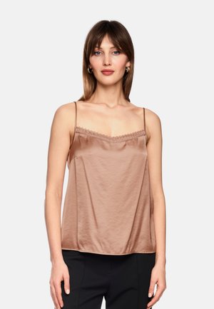 Junge Frau mit glatten braunen Haaren trägt ein ärmelloses beiges Satintop mit dünnen Trägern und schwarze Hose, steht vor schlichtem Hintergrund.