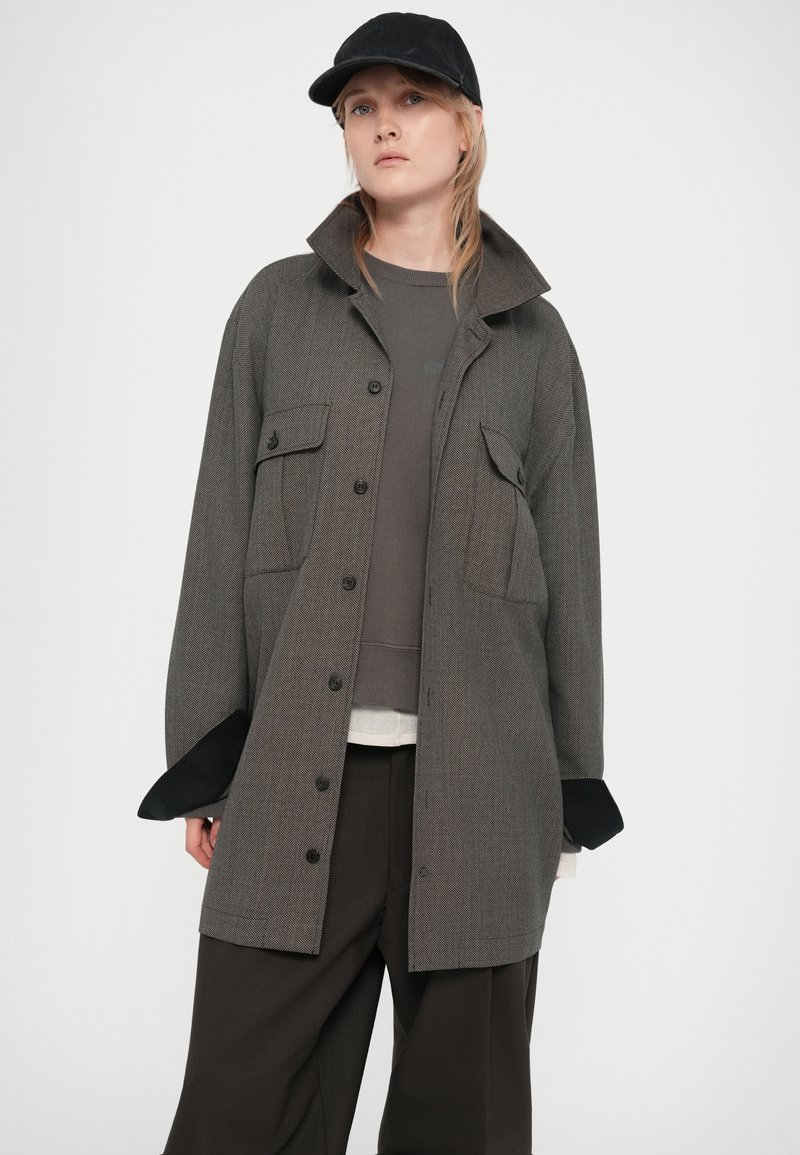 Femme portant une veste boutonnée gris oversize par-dessus un sweat gris et un pantalon large noir, avec une casquette noire, devant un fond uni.