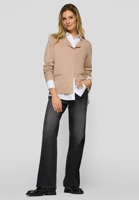 Beiger Cardigan mit Schalkragen, Knopfverschluss und zwei Taschen, über ein weißes Hemd getragen. Dunkle ausgestellte Jeans und schwarze Schuhe.