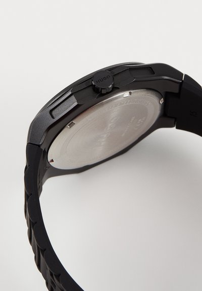 Montre en silicone noire avec un bracelet texturé, un boîtier rond plat, un dos en acier inoxydable et une couronne proéminente étiquetée "HUGO".