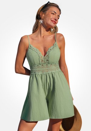 Kobieta w jasnooliwkowej sukience bez rękawów z koronkowymi detalami, trzymająca słomkowy kapelusz, uśmiechająca się, z okularami przeciwsłonecznymi na głowie na jednolitym tle.