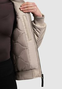 Hellbeige gesteppte Bomberjacke mit gerippten Bündchen. Mit durchgehendem Reißverschluss, einer Innentasche und einem strukturierten Futter. Glatte Außenseite.