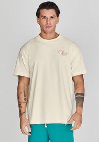 Camiseta de manga corta color marfil con un logo sutil en suave rosa en el lado izquierdo del pecho. Hecha de una tela suave, con un corte relajado y cuello redondo.