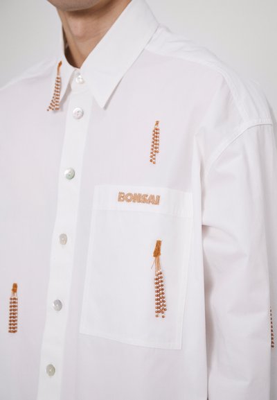 Hvid skjorte med knaplukning, der har kontrasterende brune broderidetaljer samt en lomme med "BONSai" broderet på.