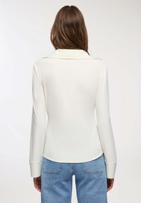 Donna con una blusa crema a maniche lunghe e jeans azzurri chiari, in piedi di spalle su uno sfondo bianco.
