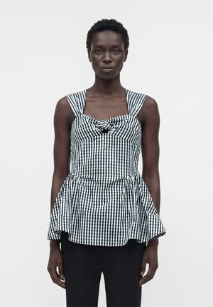 Nő fekete-fehér kockás ujjatlan felsőt visel, elöl csomózott, bővülő aljjal, fekete nadrággal párosítva, fehér háttér előtt áll.