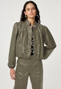 Vrouw met lang, donker haar, gekleed in een olijfgroene leren jas en broek, met daaronder een gestreept shirt en zilveren ringoorbellen, die rustig staat.