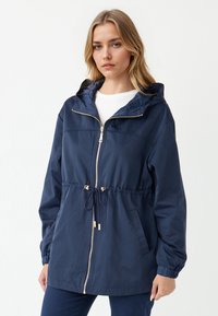 Giacca con cappuccio blu navy realizzata in materiale leggero, con vita regolabile tramite cordino, chiusura con zip e tasche laterali.