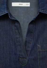 Chemise en denim bleu foncé avec col, encolure en V et poche poitrine. Présente des coutures jaunes contrastantes sur les bords et les coutures.
