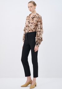 Brown spotted sheer blouse with ruffled sleeves, paired with tailored black pants and beige heeled shoes, set against a white background.