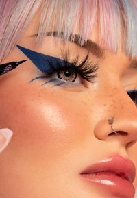 Primo piano di un occhio con eyeliner blu e un'ala drammatica, ciglia lunghe e un anello al naso a forma di farfalla argentata, su una pelle liscia e chiara.