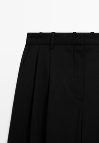 Schwarze High-Waist-Hose mit Falten, seitlichen Taschen und Gürtelschlaufen, von vorne oben auf weißem Hintergrund dargestellt.