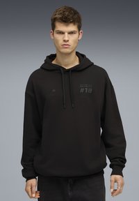 Jonge man in een zwart BMW Motorsport hoodie met de capuchon naar beneden, staand tegen een effen grijs verloopachtergrond.