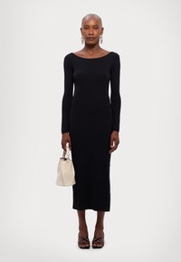 Abito lungo nero con maniche a tre quarti, scollatura a barca, silhouette aderente e tessuto morbido. Abbinato a una borsa bianca e décolleté nere con cinturini.