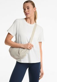 Borsa a spalla leggera di colore crema con una finitura testurizzata, portata da una persona che indossa una maglietta bianca aderente e leggings blu navy.