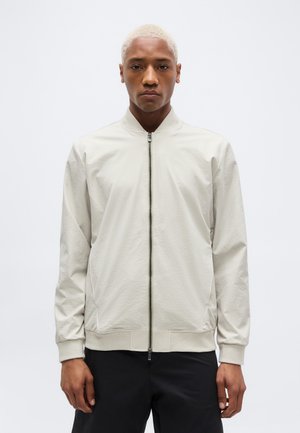 Mann mit kurzen blonden Haaren, der eine hellbeige Jacke mit Reißverschluss und schwarze Hose trägt, vor einem schlichten weißen Hintergrund stehend.