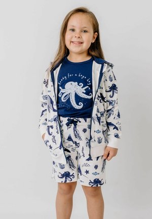 Smiling young girl wearing a navy octopus t-shirt with "ready for a huge hug" text, and matching white hoodie and shorts with sea creatures.