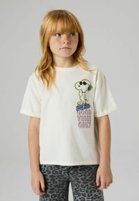 Ung rødhairet barn med fregner iført hvit Snoopy T-skjorte med teksten "Good Vibes Only" og grå bukser med leopardmønster, stående mot en ensfarget bakgrunn.