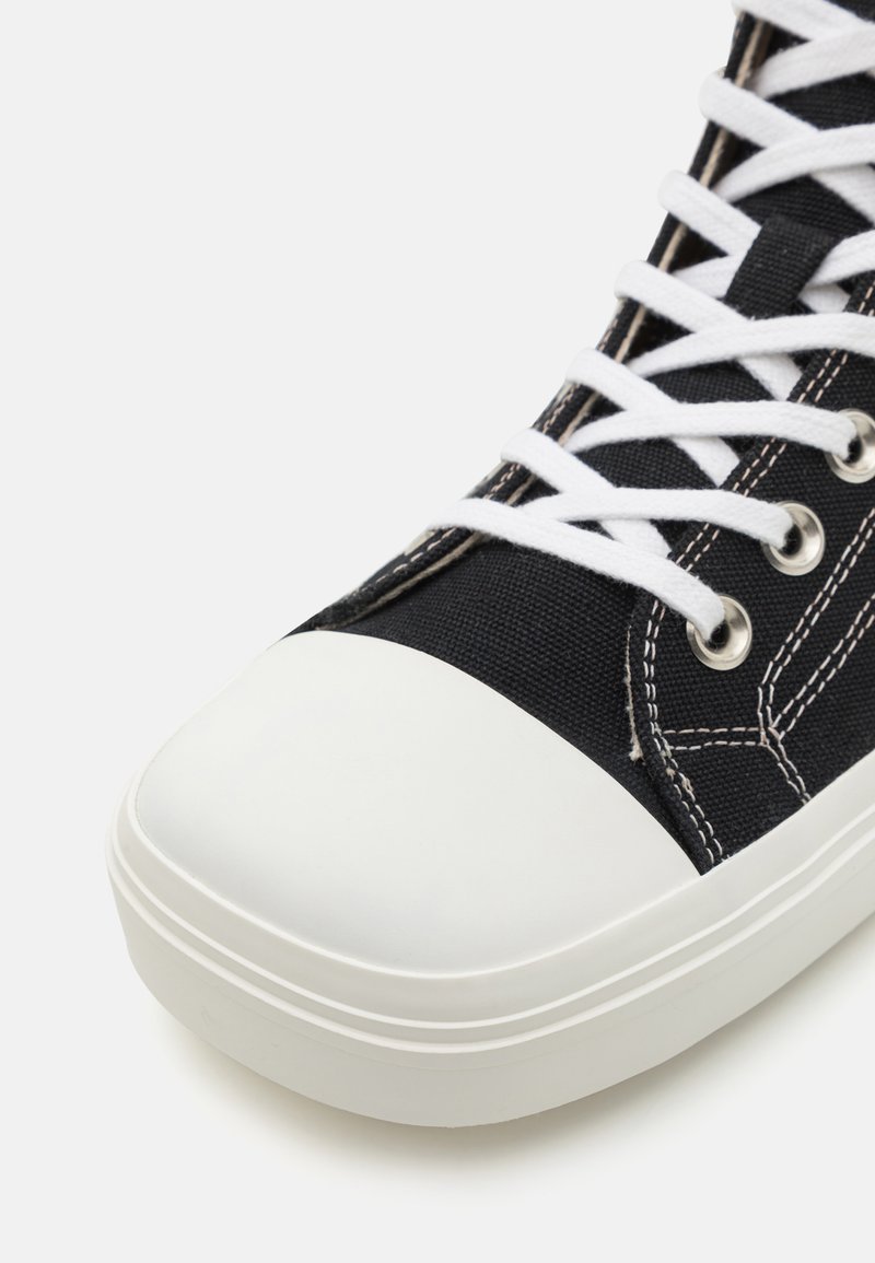 Sort lærredssneaker med hvid gummitåsok, prydet med hvide snørebånd og syninger samt sølvøjer. Fokuser på den øverste del af skoen.