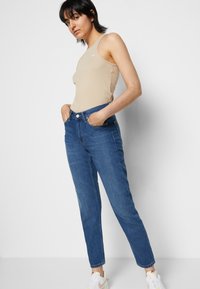 Beige ribbed sleeveless top paired with high-waisted dark blue denim jeans. Model stands with hands in pockets, wearing white sneakers.