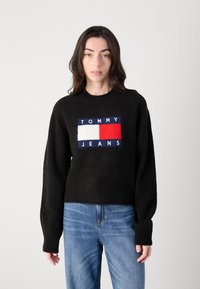 Svart strikket genser med ribbestrikkede ermer, med et sentralt logopatch i rødt, hvitt og blått. Brukt med blå jeans.