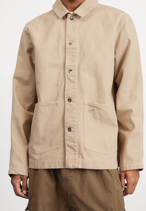 Hellbraune Knopfleistenjacke mit zwei großen Fronttaschen, getragen von einer Person, kombiniert mit dunkelbrauner Hose.