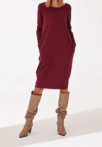 Kobieta ubrana w burgundową midi sukienkę z długim rękawem i kieszeniami oraz luźne beżowe wysokie botki na obcasie, stojąca na białym tle.