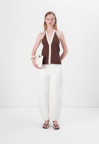 Hnedý rebrovaný halter top s krémovým lemovaním a detailmi gombíkov, v kombinácii s voľnými bielymi nohavicami a čiernymi sandálmi na pásiky, so bielou taškou v ruke.