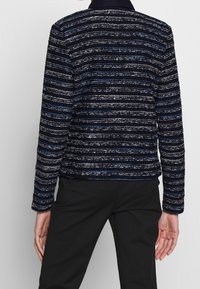 Femme portant une veste texturée à rayures bleu marine et noir et un pantalon noir, vue de dos sur un fond uni.