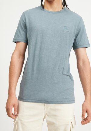 Mand iført en lyseblå T-shirt med korte ærmer og et lille firkantet logo på brystet, kombineret med cremefarvede cargobukser.