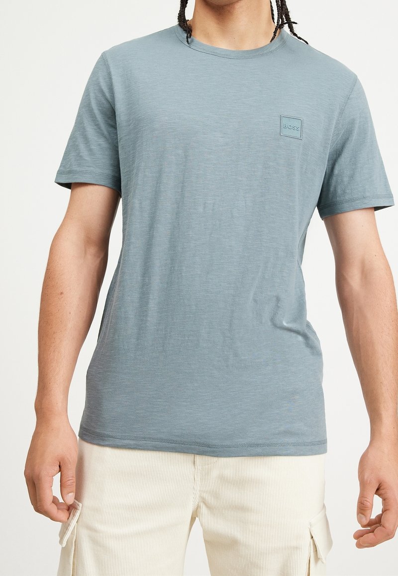 Homme portant un T-shirt à manches courtes bleu clair avec un petit logo carré sur la poitrine, associé à un short cargo crème.