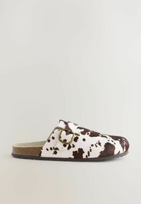 Zueco blanco de tipo slip-on con un patrón de piel de vaca marrón, que presenta un acento de hebilla metálica y una suela texturizada marrón.