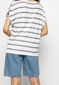 Persona vestida con una camiseta de rayas blancas y negras de manga corta y un pantalón corto de mezclilla azul holgado, de pie contra un fondo claro y liso.