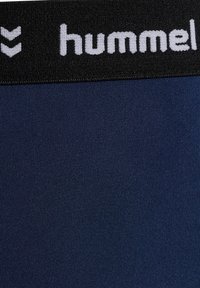 Granatowe legginsy sportowe z czarnym elastycznym pasem w talii, z białym logo "hummel" i wzorem chevron. Gładki, elastyczny materiał.