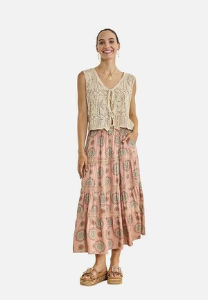 Mujer sonriente con top sin mangas beige de crochet atado al frente, falda larga rosa con flores, sandalias con plataforma y joyería dorada.